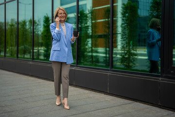 Obraz premium Mature businesswoman walking, talking on smartphone, holding coffee