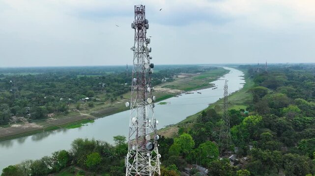 Aerial view of a tall telecommunication tower and a secondary mast beside a winding river with birds flying over lush green trees and rural fields Bangladesh.