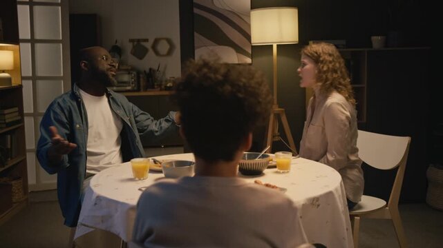 Back view of Black teenage boy covering ears while watching parents fighting and yelling at dinner table at home, conflict in multiracial family, copy space