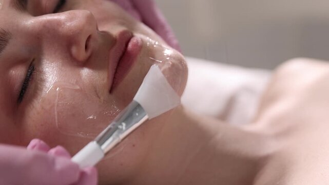 Beautician in medical gloves applying chemical peel with cosmetic spatula onto client's face indoors, closeup