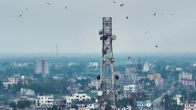 Aerial view of a tall telecommunications tower with antennas and satellite dishes overlooking a dense urban area with birds flying in an overcast sky.