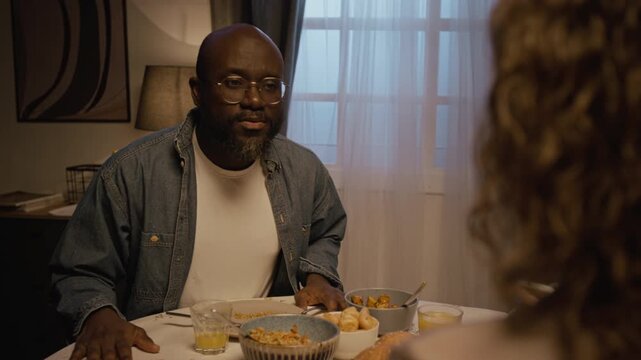 Candid over shoulder shot of adult couple fighting while sitting at dinner table at home with focus on Black man talking, scene lit by low evening lighting