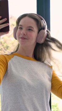 Vertical video: Raising phone teen in yellow sleeves filming selfie at window, fixing pink headset
