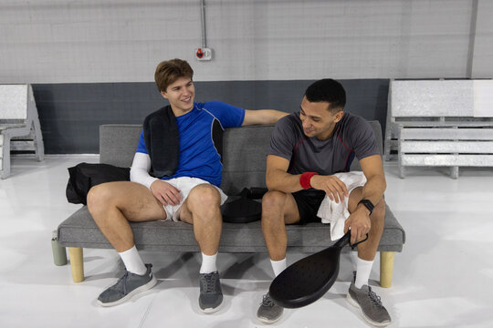 Male teammates in sportswear sitting on gray sofa in gym lounge holding perforated paddle and towel