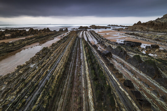 View of Playa de Sopelana featuring unique flysch rock formations under a dramatic cloudy sky in Vizcaya, Pais Vasco, Spain.