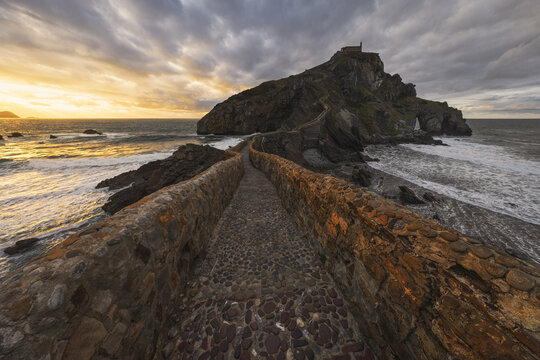 View of the stone bridge leading to the hermitage on a rocky island under a dramatic sunset sky with crashing waves Gaztelugatxeko Doniene, Euskadi, Spain.