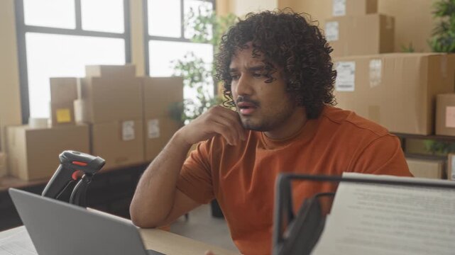Man in orange shirt holding document and hand on cheek beside laptop and barcode scanner in building; small business concentration.