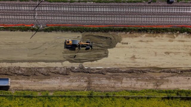Construction Site Activity: An aerial view captures a loader working on a muddy construction site, with railway tracks and green fields nearby, suggesting ongoing development and infrastructure work.
