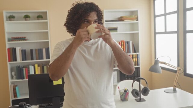 Man holds coffee cup with both hands and looks up at window in a building filled with bookshelves and desk; calm reflection.