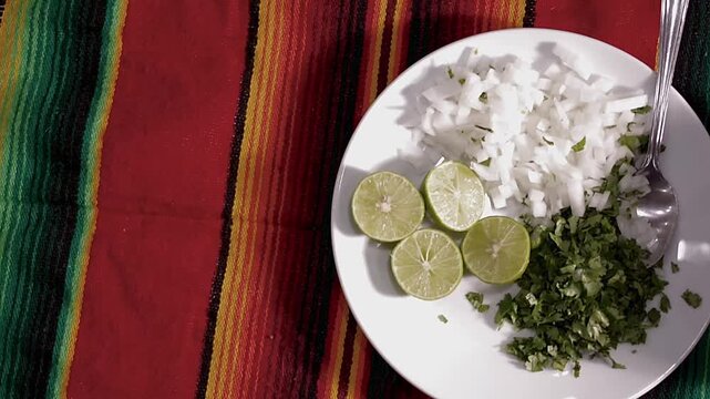 Plato blanco con limones cortados cebolla y cilantro picados sobre una mesa con mantel de colores para complementar comida Mexicana. Vista Cenital