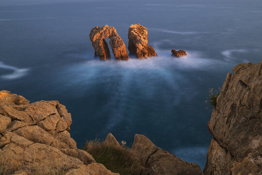 View of the Urro del Manzano sea arch and rugged rock formations surrounded by misty long exposure waves during sunset Liencres, Cantabria, Spain.