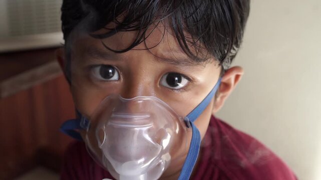 Close up of sick Asian young boy with damp hair looking at camera while wearing nebulizer mask with visible mist, unwell expression during respiratory treatment at home