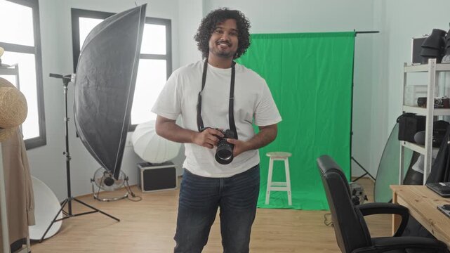 Man photographer holding camera with strap in studio, smiling and aiming lens, wearing white t shirt and jeans; creative confidence.