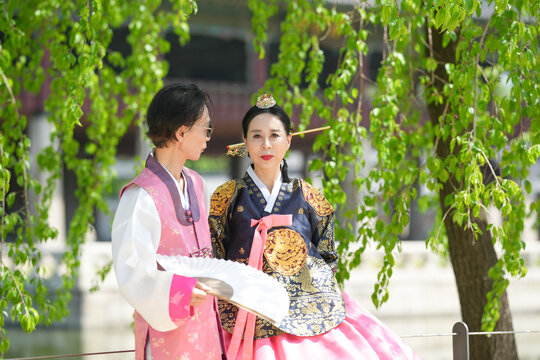 Lush green branches frame two traditional Korean figures. Woman in ornate court dress sits gracefully beside her companion. Her partner, dressed in soft pink and white, holds a ceremonial fan