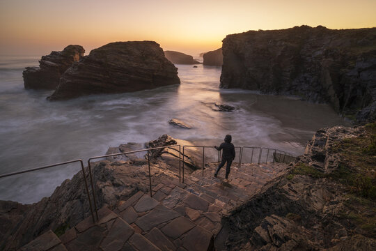 View of Praia das Catedrais with a person standing on stone stairs overlooking dramatic rock formations and sea arches during a golden sunset in Lugo, Lugo, Spain.