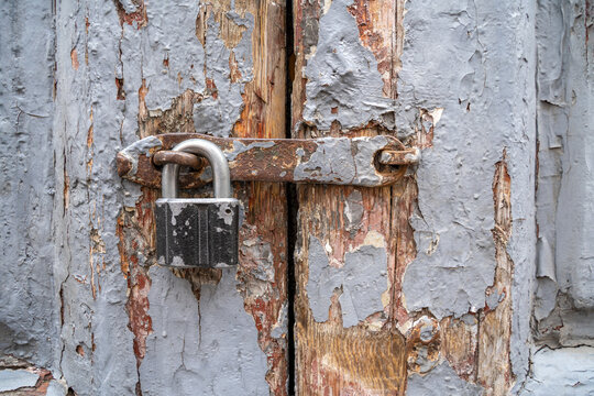 Old rusty metal padlock hanging on weathered wooden door with heavily peeling blue paint, abandoned security concept, grungy texture detail.