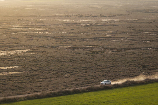 View of a white car driving on a dusty road through the arid landscape of Bardenas Reales with green fields in the foreground Navarre, Navarre, Spain.