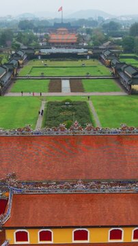 Vertical screen Aerial Axis View Of Hue Citadel Ceremonial Lawn And Central Pathway Leading To Flagdraped Building, Manicured Hedges And Fountain, Visitors Appear As Small Figures On Wet