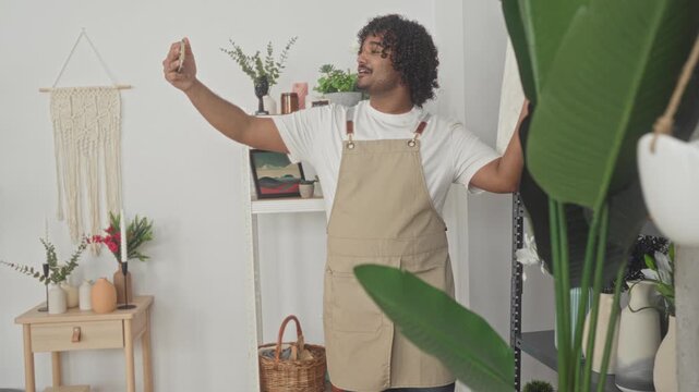 Man holding smartphone in hand taking selfie while wearing florist apron in studio surrounded by plants vases shelf and wicker basket; serenity.