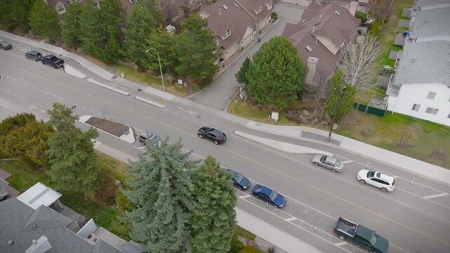 Overhead shot of a black SUV driving on a quiet road through a multi-unit neighbourhood on a cloudy day. Tree-lined street features bike lanes, pedestrian sidewalks and several parked cars. 