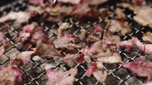 Close-up of thin slices of marbled beef grilling on a traditional charcoal barbecue net. Tongs are seen flipping the meat over glowing red embers in a Japanese yakiniku style restaurant setting.