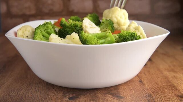 Tazon blanco con vegetales brocoli, zanahoria y coliflor cocidos sobre una mesa de madera y pared de ladrillos al fondo.
