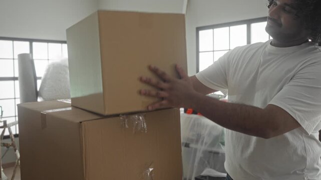 Man stacks cardboard boxes, stands with arms crossed and smiles inside a building room filled with moving supplies; moving day pride.