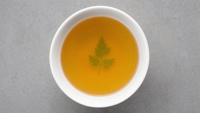 A single bowl of golden consomme with a delicate herb leaf, minimalist and pure.