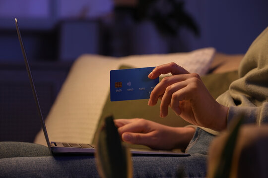 Young woman with credit card using laptop for online shopping on sofa in evening at home, closeup