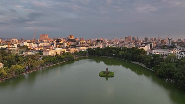 Wide aerial video panorama of Hoan Kiem Lake featuring Turtle Tower, calm reflective water, and dense Hanoi urban skyline at sunset.