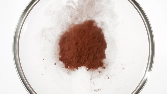 Top view macro shot of a foggy cloud of brown dust dispersing and flowing in a petri dish over a pile of powder against a clean white background