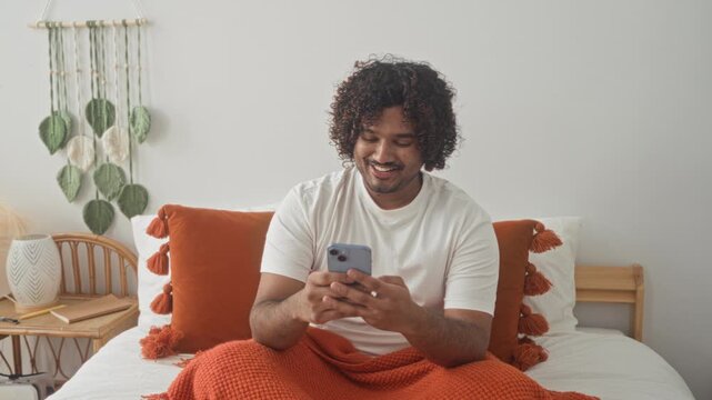 Man holds smartphone at arm's length taking a selfie on bed in studio with orange pillows, blanket and macrame wall hanging; selfcare contentment.