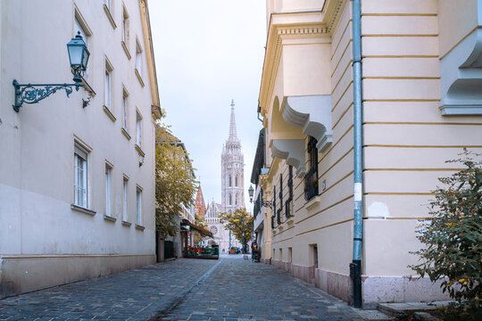 Matthias church spire rising above the quaint cobblestone street t&oacute;th &aacute;rp&aacute;d s&eacute;t&aacute;ny in budapest castle hill, historic european architecture and autumn trees
