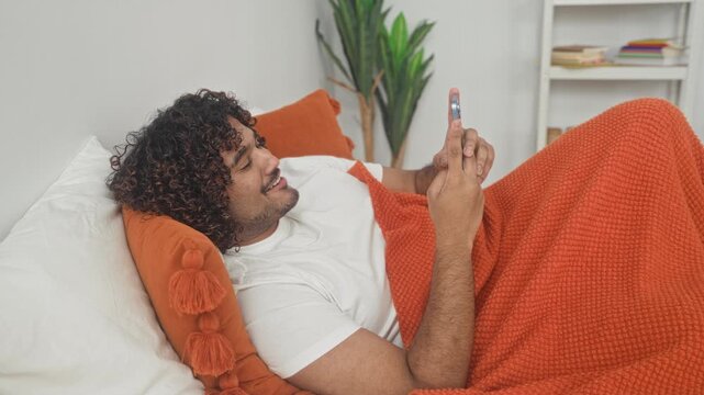 Man holding smartphone with both hands while lying on bed in studio set with orange knit blanket, tassel pillow and indoor plant visible; serenity.