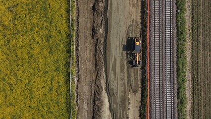 Fototapeta premium Railway Construction Aerial View: An aerial perspective reveals a construction site next to a railway track, where heavy machinery operates on bare earth adjacent to a field of yellow crops.