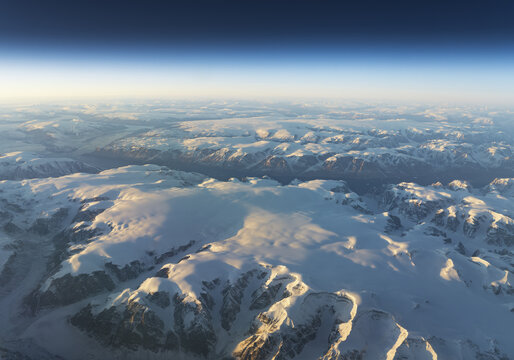 Aerial view of the vast snow-covered mountain ranges and massive glacial ice sheets under a clear blue sky during golden hour Greenland.