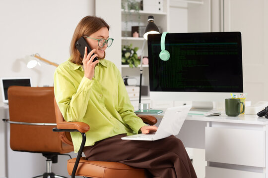 Happy female programmer with laptop and computer talking by mobile phone in office