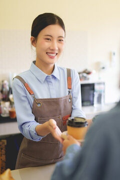 Asian female barista smiling serving takeaway coffee cup to customer at cafe counter wearing apron providing friendly service at coffee shop business