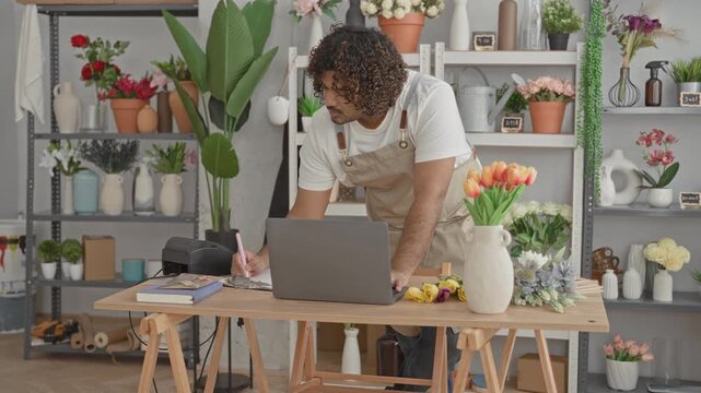 Man florist in apron leans over laptop with hands on table arranging tulips in building;friendly service.