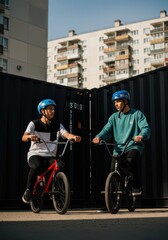 Fototapeta premium Two Young Men on BMX Bikes Talking in Urban Setting with Shipping Containers and Apartment Buildings Background