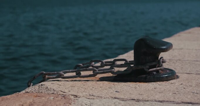 Black mooring bollard with metal chain on a marina pier beside seawater.