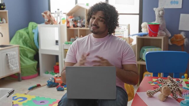 Man holds stuffed giraffe in right hand while typing on laptop among colorful kindergarten toys in building; playful learning warmth.