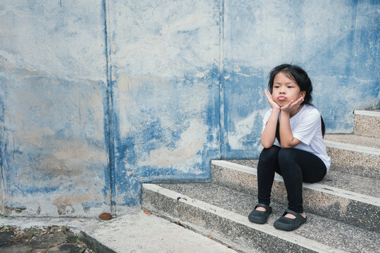 Portrait of a sad Asian little girl sulking and sitting on steps against a grunge blue wall backdrop, showcasing childhood emotions, lonely feelings, and psychological expression concepts.