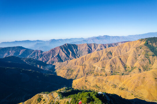 Aerial view of the rolling Himalayan foothills with winding mountain roads and lush ridges under a clear blue sky in Chakrata, Uttarakhand, India.