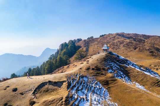 Aerial view of a small mountain temple on a ridge with a person walking along a path and snow-covered slopes in Chakrata, Uttarakhand, India.