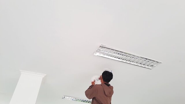 PAMEKASAN, INDONESIA - JANUARY 06, 2026: Professional network technician upgrading internet connectivity by installing a new high-speed Wi-Fi access point on a minimalist room ceiling. The technician 