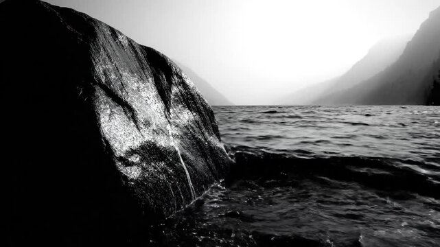 Black and white scenic video of water lapping against a large, dark, glistening rock with mountains in the background