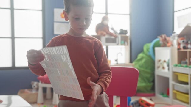 Boy holds an alphabet sticker sheet with both hands and studies the letters while standing in a building playroom; focused play.
