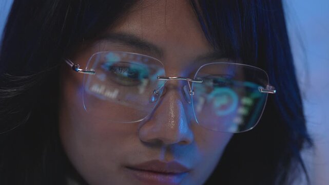 Close up portrait of a young Asian female software developer wearing rimless glasses with vivid blue code reflection in the lenses while focused on her bright computer screen