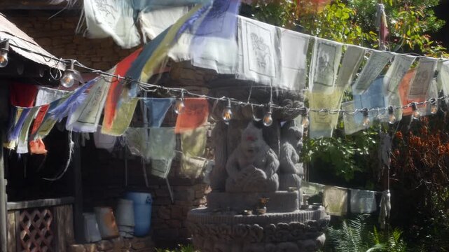 Prayer flags flutter in the wind above a stone deity statue and string lights in a Tibetan shrine courtyard. Sunlit fabric, carved sculpture, and rustic stone walls create a layered outdoor religious